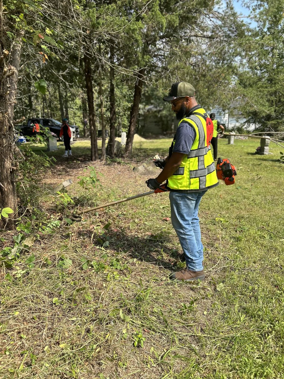 Volunteer using a string trimmer to clear brush along the tree line at Snow Hill Cemetery